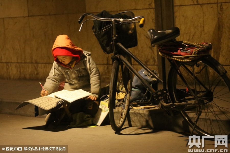 鄭州中原路京廣鐵路大橋下涵洞的馬路牙子邊，一位戴著紅色辣椒帽的小男孩趴在一個小馬扎上，上面鋪著一張車墊，在微弱的燈光下寫作業(yè)