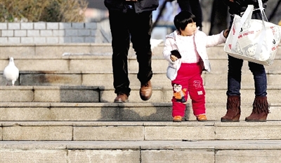 昨天中午，一女孩拉著家長的手在紫荊山公園曬太陽，氣溫升高，孩子的棉衣解開了扣子。